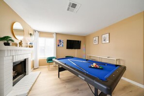 Une table de billard recouverte d’un drap bleu, une cheminée avec des carreaux blancs et un fauteuil vert.