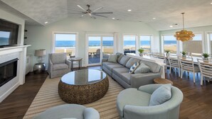 Un salon spacieux doté d’une cheminée, de sièges confortables, d’un coin repas et de grandes fenêtres offrant une vue sur la plage.