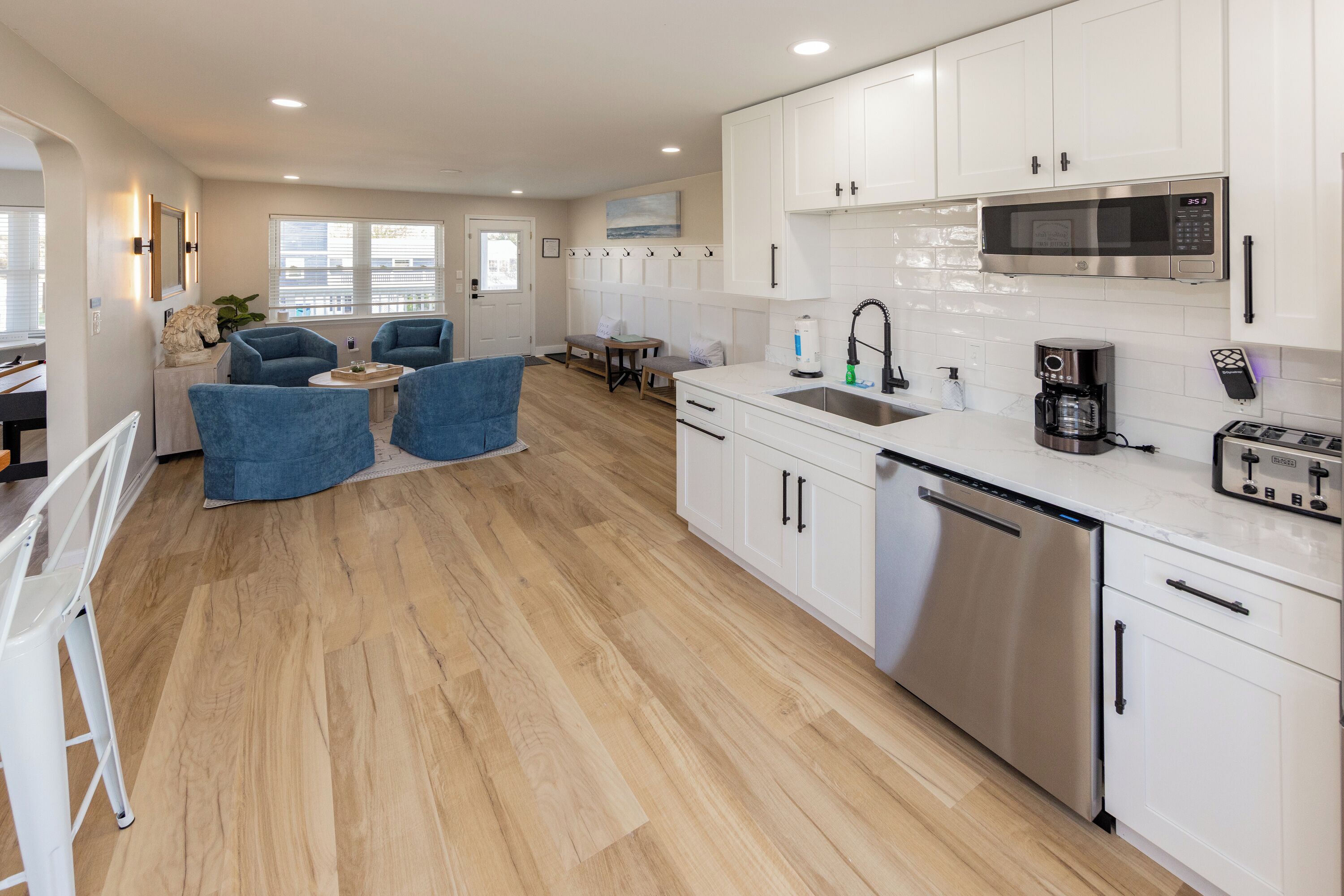 Cocina moderna con armarios blancos, electrodomésticos de acero inoxidable y suelo de madera. Hay una zona de estar con sillas azules y una mesita.
