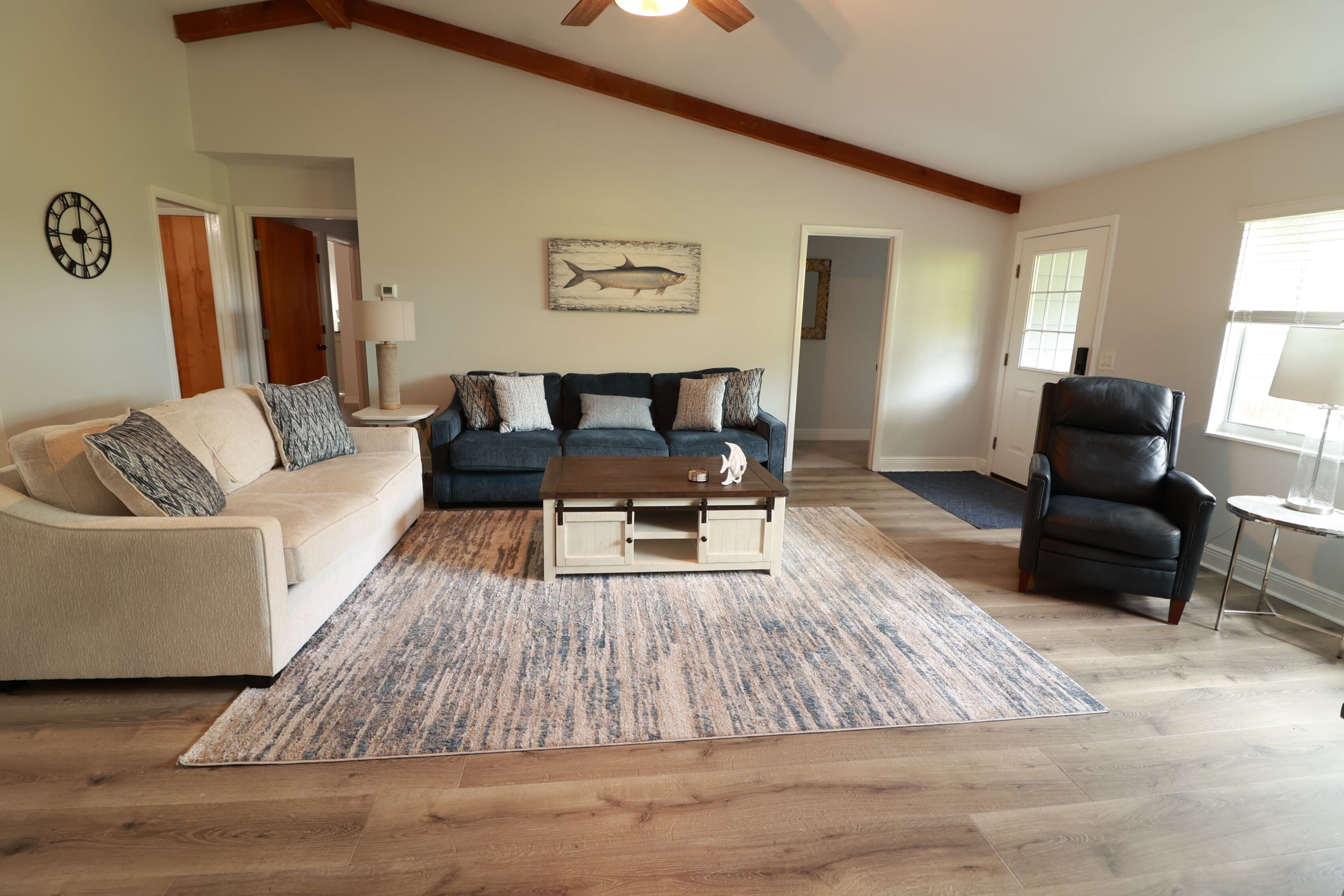 Ein Wohnzimmer mit einem beigefarbenen Sofa, einem blauen Sofa, einem Holztisch und einem schwarzen Ledersessel. An der Wand hängt ein Fischgemälde und eine Uhr.