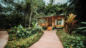 A wooden cabin with a porch, surrounded by lush greenery and a concrete pathway. - HOSTERIA KUMBHA MELA (Mindo)