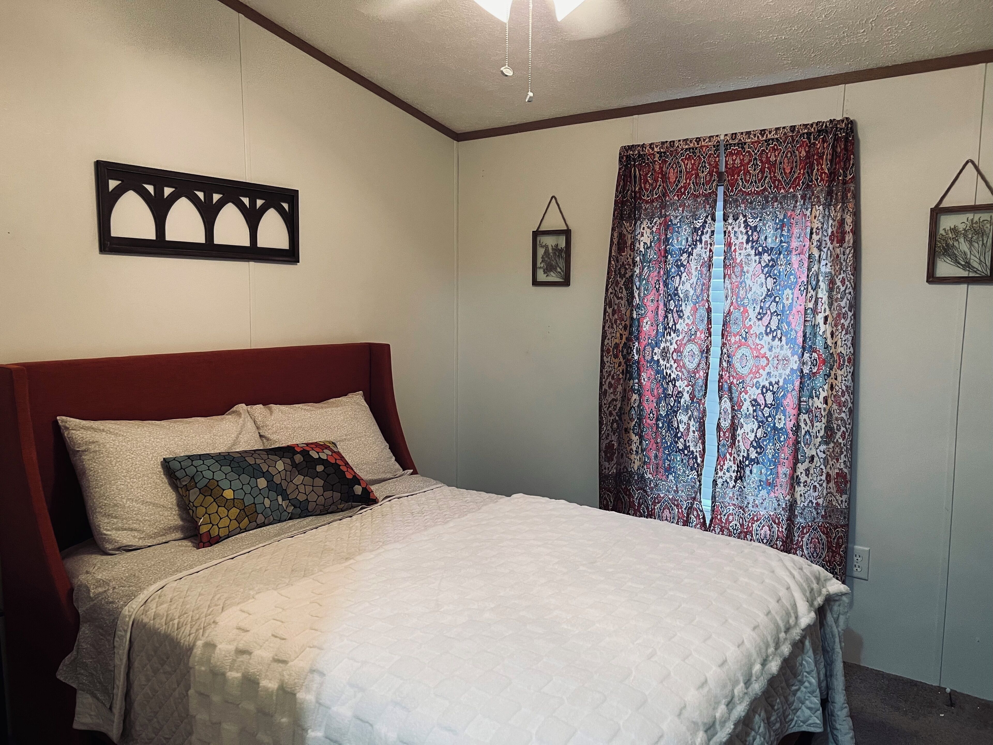 A bedroom with a bed, a headboard, a patterned curtain, and a wall-mounted picture frame.