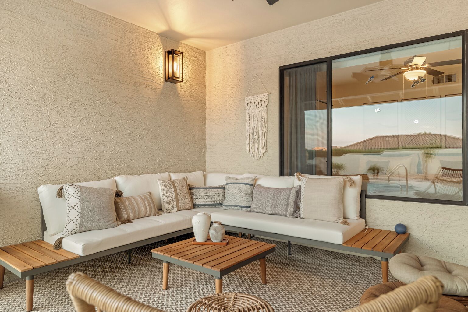 A modern living room with a white sectional sofa, wooden coffee table, and wicker chairs. A large window offers a view of a pool and desert landscape.