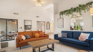 Un salón espacioso con un sofá marrón, un sofá azul y una mesa de centro de madera. Hay plantas en macetas en el alféizar de la ventana y un espejo grande en la pared.