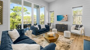 A modern living room with a dark blue sofa, white armchairs, and a wooden coffee table. Large windows offer a view of the outdoors. - 7-bedroom/ 7-bathroom house; Private pool; Golf cart included (Santa Rosa Beach)