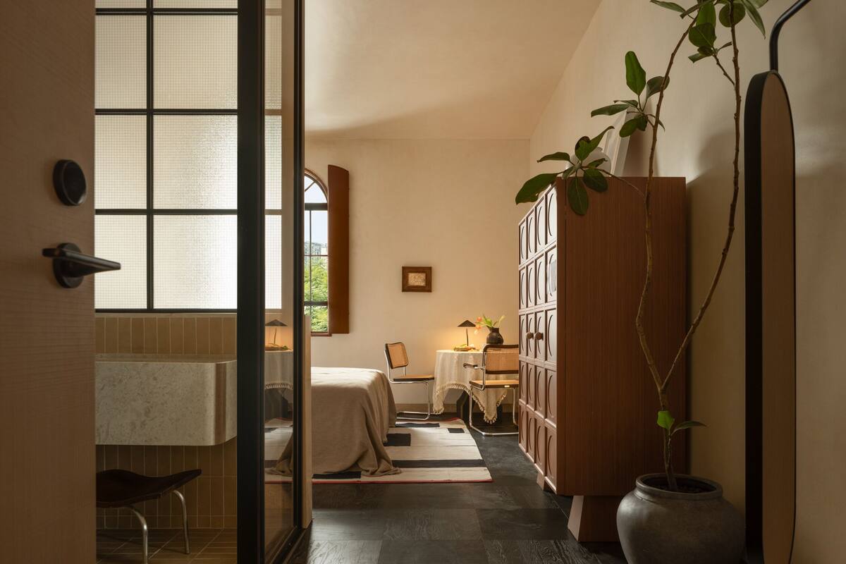 A hotel room with a bed, a desk, a chair, a bookshelf, and a potted plant.