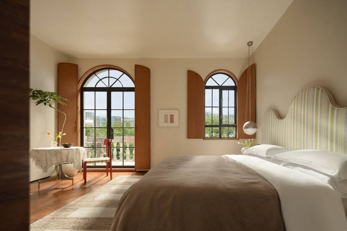 A bedroom with a large bed, two windows with wooden shutters, a small table with a chair, and a view of greenery outside.