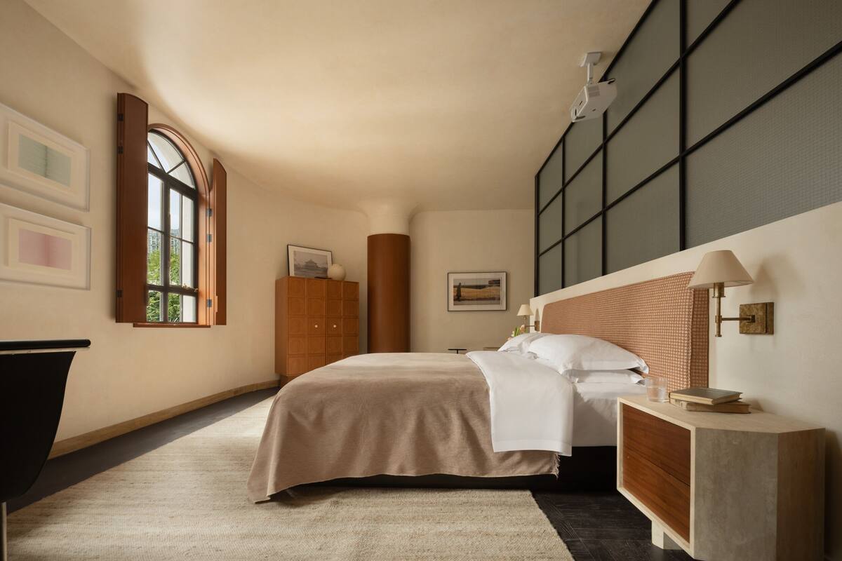 A bedroom with a large bed, a nightstand, a wooden wardrobe, a window with a view, and a wall-mounted projector.
