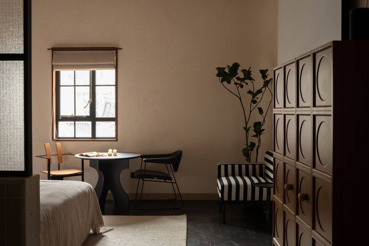A room with a bed, a round table with chairs, a window with a view of buildings, and a plant.