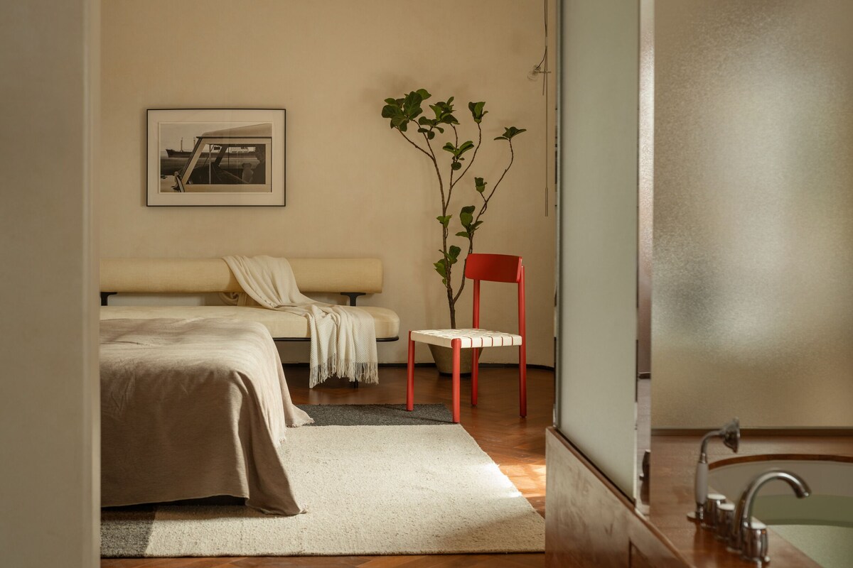 A bedroom with a bed, a chair, a plant, and a framed picture on the wall.