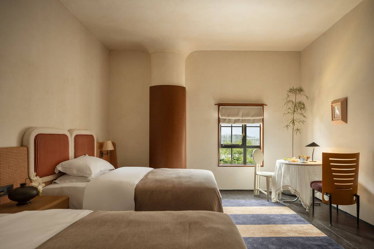 A hotel room with two beds, a window with a view, a small table with a chair, and a plant.