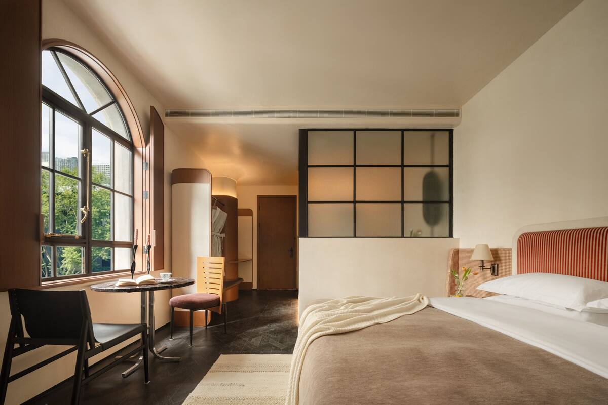 A hotel room with a large window, a bed, a desk with a chair, and a small table.