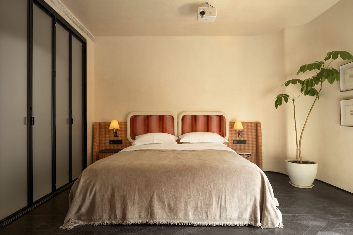 A neatly made bed with a beige blanket, two bedside lamps, a potted plant, and a sliding door on the left.
