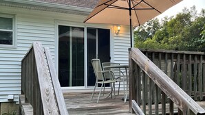 Une terrasse en bois avec une table et des chaises, un grand parasol et un escalier menant à une maison dotée d’une porte-fenêtre coulissante.