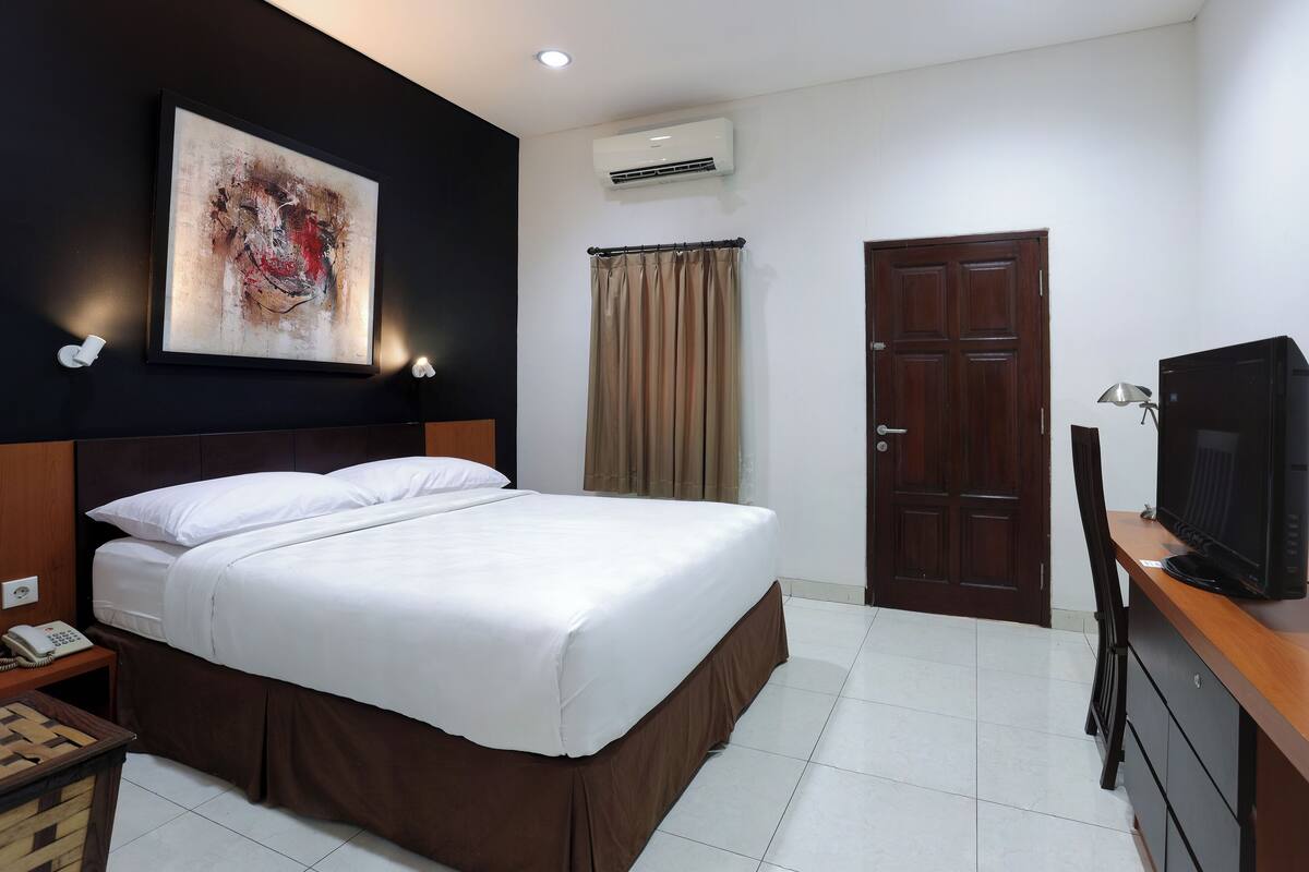 A hotel room with a large bed, a desk with a television, a wardrobe, an air conditioning unit, and a painting on the wall.
