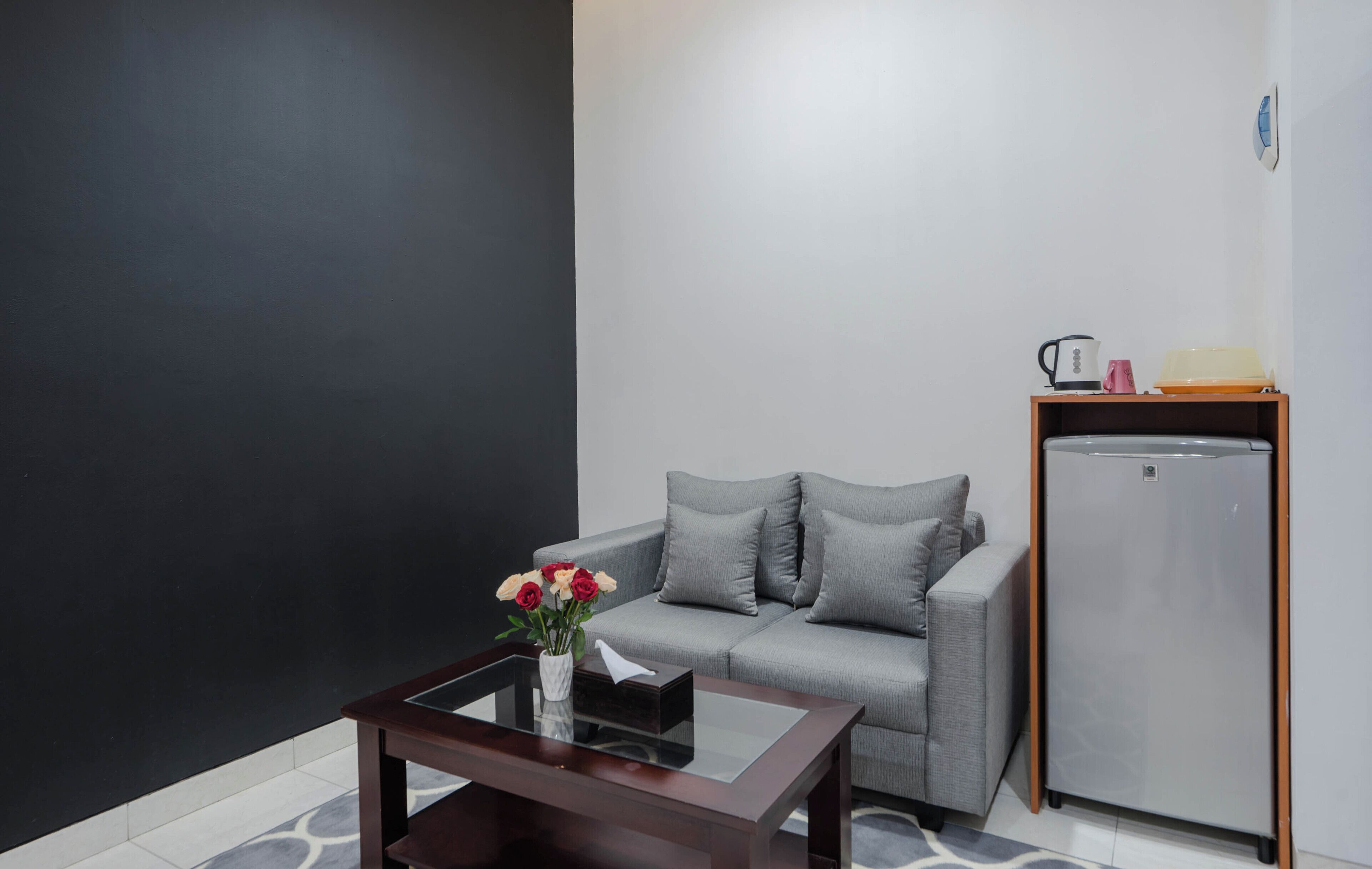 A modern living room with a grey sofa, a small table with a vase of flowers, a white wall, and a refrigerator.