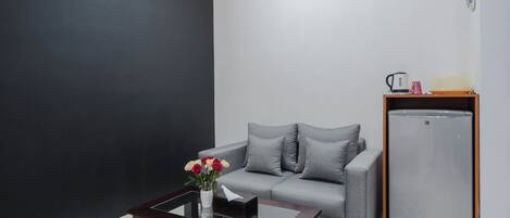 Una sala de estar moderna con un sofá gris, una mesita con un florero y una pared blanca, además de un refrigerador.