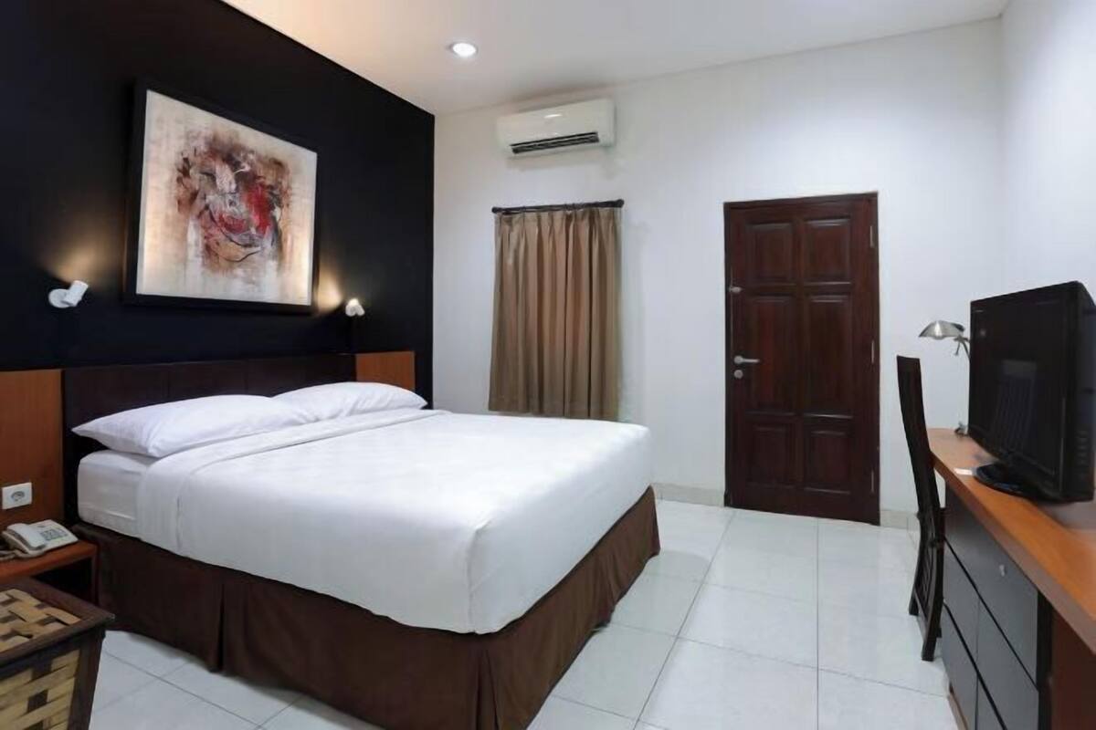 A hotel room with a bed, a television, a desk, a chair, an air conditioning unit, a door, and a painting on the wall.