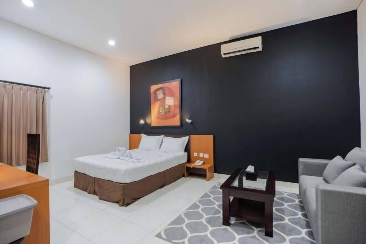 A hotel room with a bed, a desk, a chair, a sofa, a painting on the wall, and an air conditioning unit.