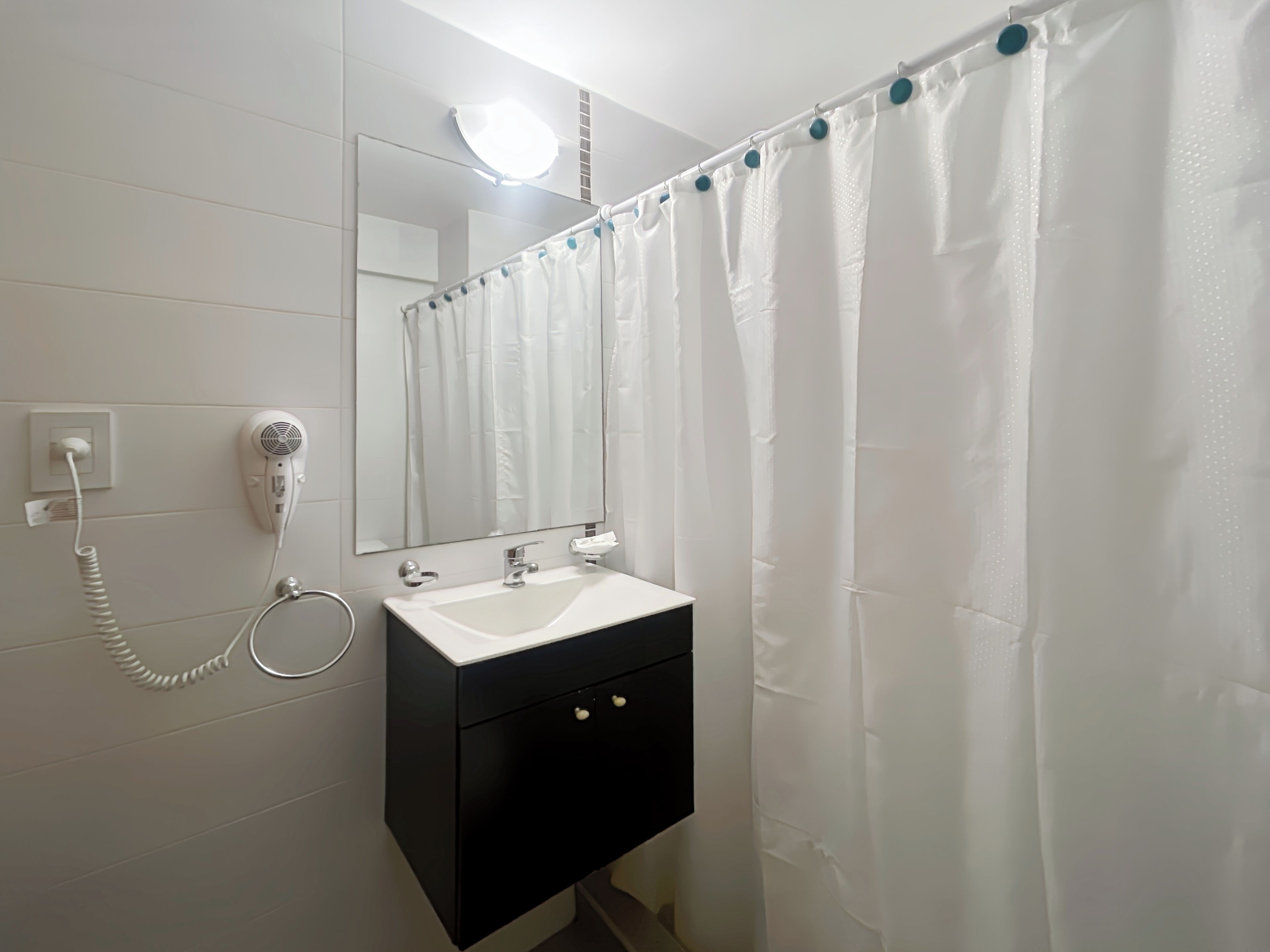 Un baño con lavamanos blanco, secador de pelo, espejo y cortina de ducha.