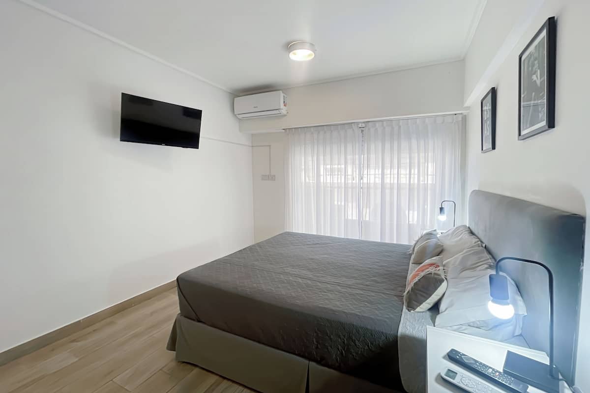 A modern bedroom with a bed, a bedside table with a lamp, a mounted TV, and an air conditioning unit.
