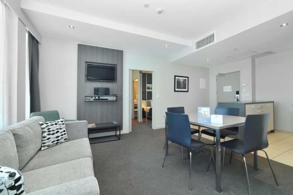 A modern living room with a grey sofa, a dining table with chairs, a flat-screen TV mounted on the wall, and a kitchen area with cabinets and a countertop.