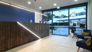 A modern reception area with a wooden reception desk labeled "QUEST," a glass door leading outside, and a seating area with chairs. - Quest Highbrook (Auckland)