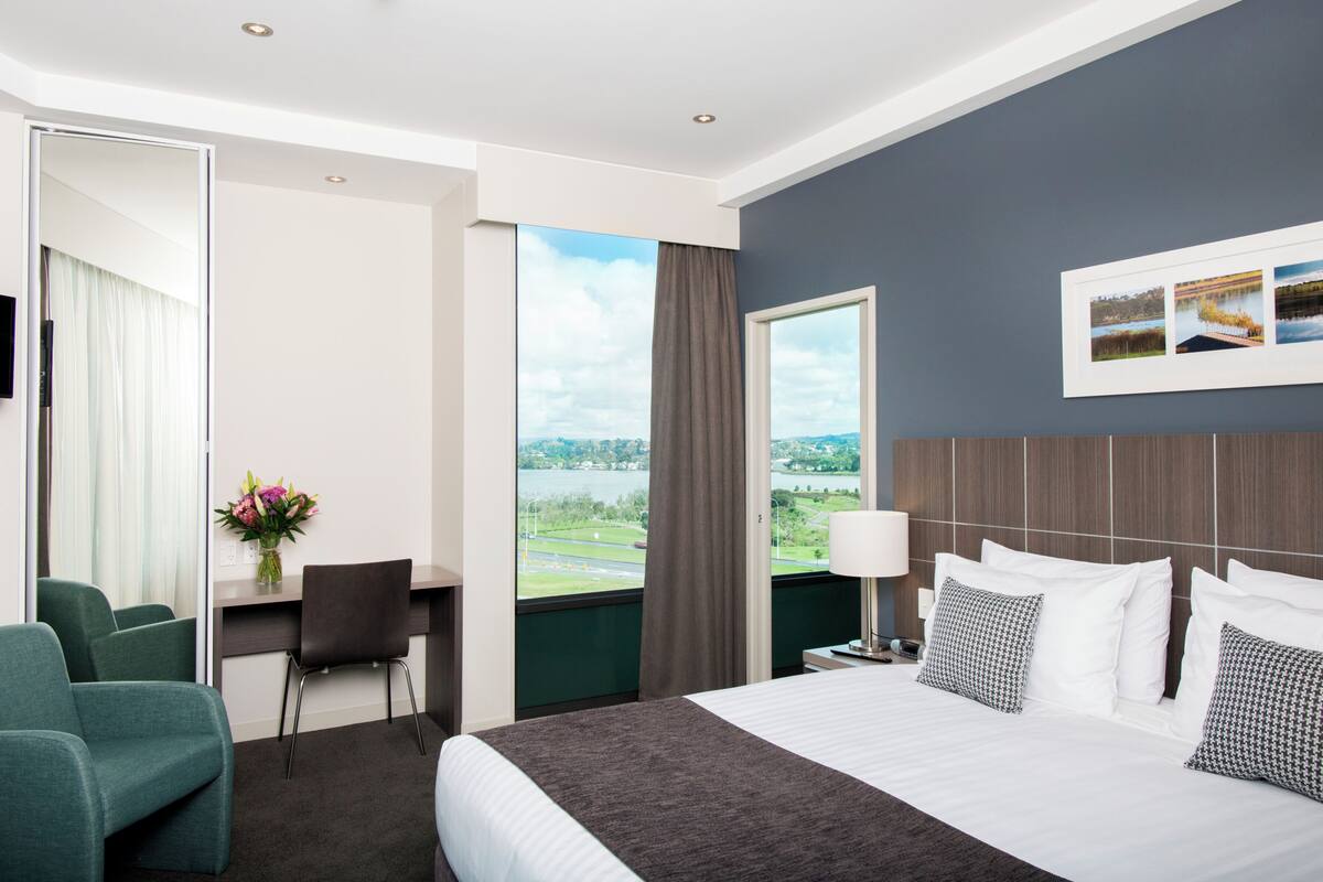 Habitación de hotel con cama, escritorio, silla, televisor, ventana con vistas y un cuadro colgado en la pared.