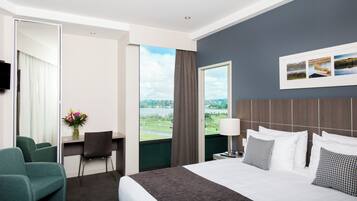 A hotel room with a bed, a desk, a chair, a TV, a window with a view, and a wall-mounted picture.