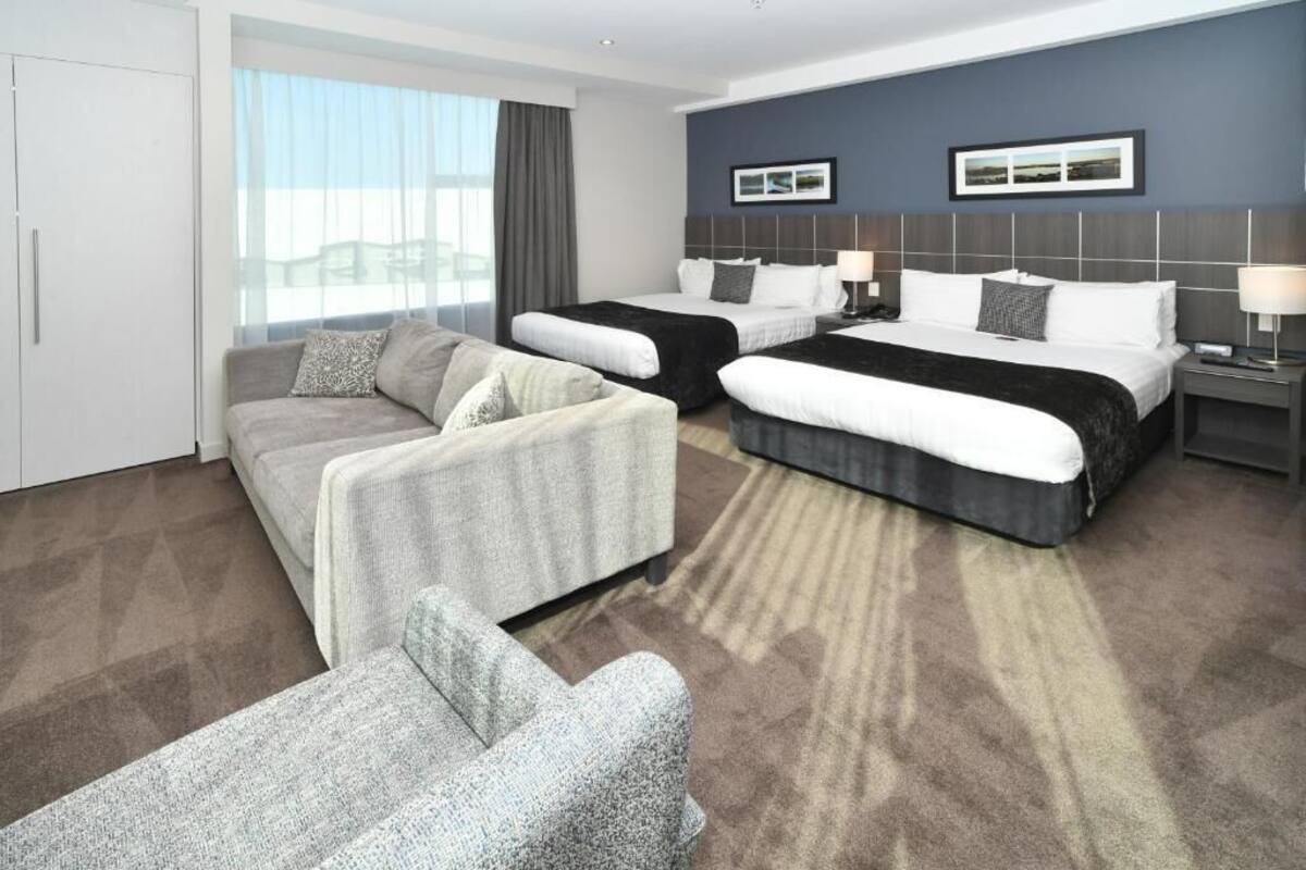 Habitación de hotel con dos camas, un sofá, una silla, un ventanal grande y vista a la ciudad.