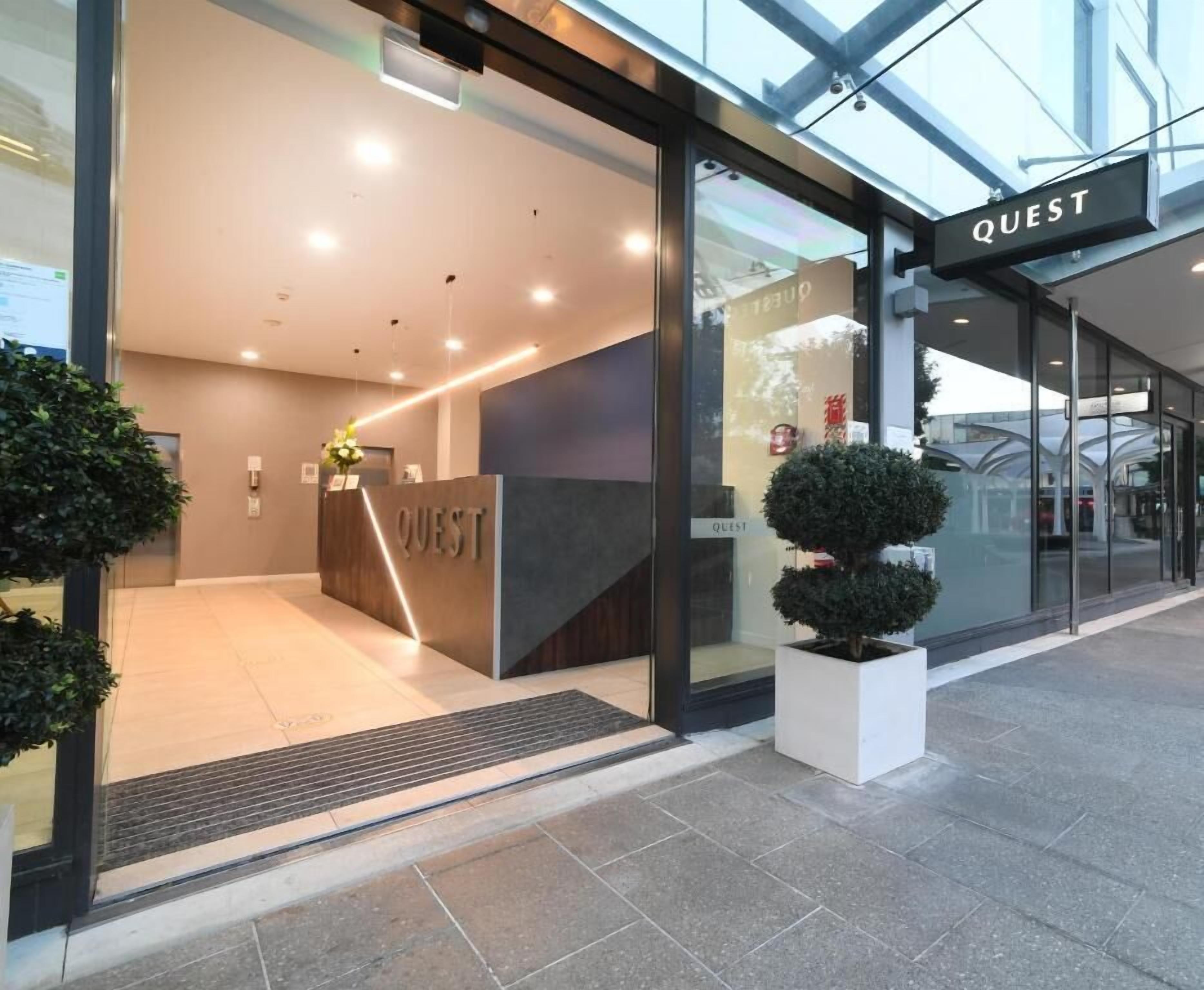 Entrada moderna de un hotel con una recepción que dice "QUEST", una puerta de vidrio y una pequeña planta en maceta afuera.