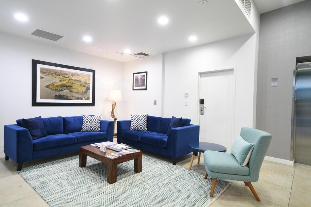 Un salon moderne avec deux canapés bleus, une table basse et un fauteuil bleu. Un tableau encadré est accroché au mur, et une porte se trouve sur le côté droit.