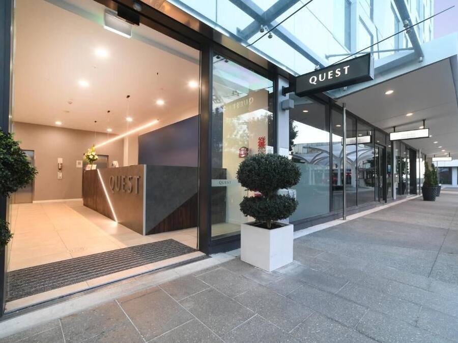 Una entrada de hotel moderna con un mostrador de recepción que dice "QUEST" y fachada de vidrio.