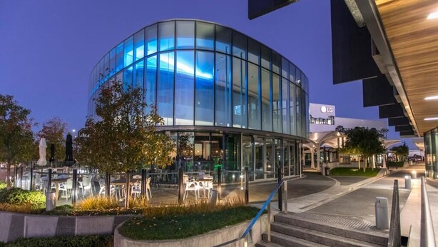 A modern building with a curved glass facade, outdoor seating area, and a staircase leading to the entrance. - Quest Highbrook (Auckland)