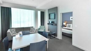 A modern hotel room with a dining area, a sofa with patterned cushions, a TV mounted on the wall, and a separate bedroom with a bed and nightstand. - Quest Highbrook (Auckland)