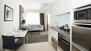 A modern kitchen with stainless steel appliances, a dining area with a table and chairs, and a living space with a sofa and curtains. - Quest Highbrook (Auckland)