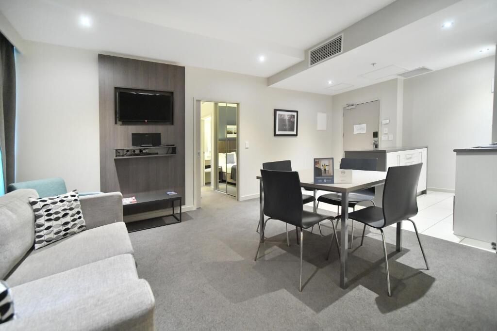 Una sala de estar moderna con un sofá gris, una mesa de comedor con cuatro sillas, un televisor de pantalla plana montado en la pared y una pequeña zona de cocina con un refrigerador y un microondas.