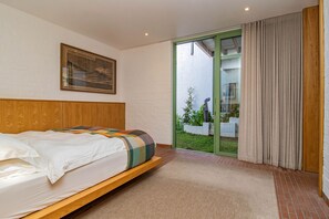 A bedroom with a bed, a painting, a sliding glass door to a balcony, and a view of a garden.