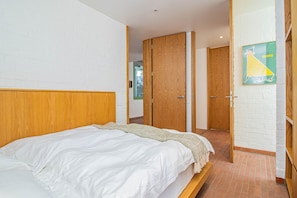 A bedroom with a bed, wooden headboard, white walls, a wooden door, and a poster on the wall.