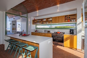 A modern kitchen with a central island, bar stools, wooden shelves, and a view through the window.