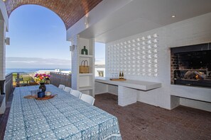 An outdoor dining area with a view of the sea, a fireplace, and a white brick wall with shelves.