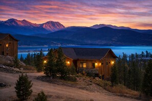 A rustic cabin with a stone foundation, surrounded by trees, overlooking a lake and mountains. - Luxe Summer Chalet: Epic Lake Views, Cinema & Gym - 8 Min to RMNP + Hot Tub (Grand Lake)