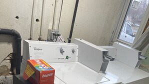 A laundry room with a Whirlpool washer and dryer, a box of Bounce detergent, and a water meter.