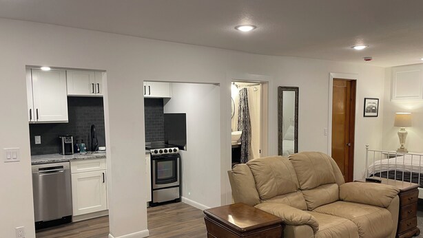 A modern living room with a beige sofa, a wooden side table, and a kitchen area with white cabinets and a stove.