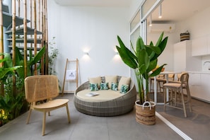 A modern living area with a round seating area, a rattan chair, a large potted plant, and a kitchen in the background.