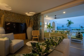 A modern living room with a stone wall, a flat-screen TV, and a balcony with a view of palm trees and mountains.
