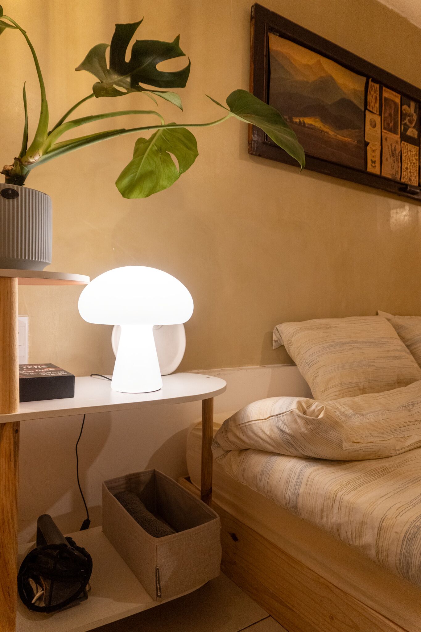 A bedroom with a bed, bedside table, lamp, plant, and a framed picture on the wall.