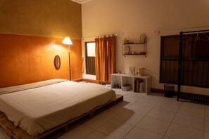 A bedroom with a bed, a nightstand, a shelf with items, a window with curtains, and a door with a handle. - THE Room (Ho Chi Minh City)