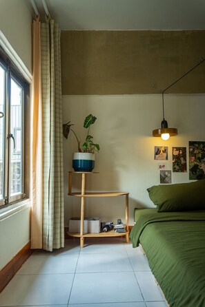 Ett sovrum med en säng, ett fönster med gardiner, en hylla med en växt och en vägg med tavlor.