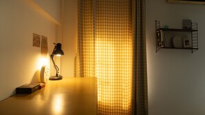 Ein Zimmer mit Schreibtisch, Lampe, einer Uhr, einem Bücherregal und einem Fenster mit kariertem Vorhang.